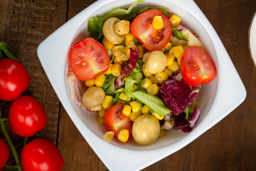 Vegetable salad with champignon corn and cherry tomato with avocado in a white plate on a wooden table.