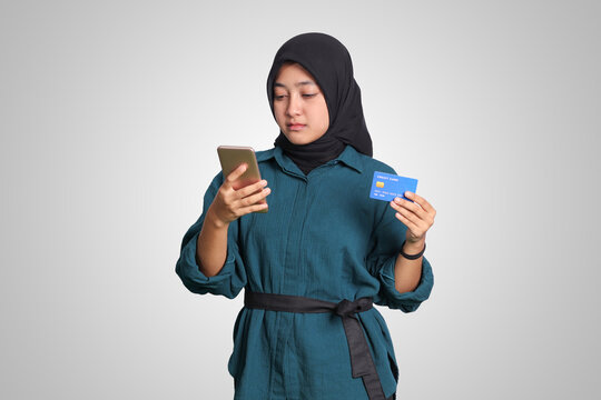 Portrait of cheerful Asian muslim woman with hijab, showing credit card while holding mobile phone. Financial and savings concept. Isolated image on white background