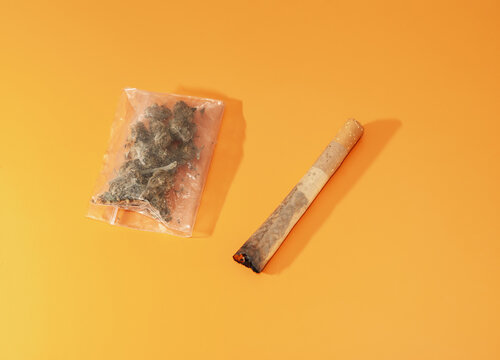 Cigarro porro y bolsita de pl&aacute;stico con marihuana en su interior sobre fondo rugoso de color naranja. Orientaci&oacute;n inclinada.