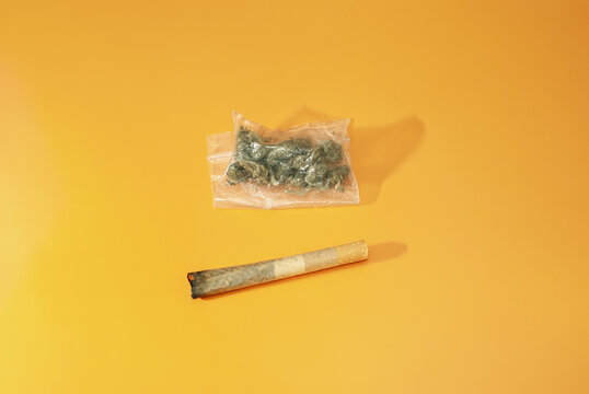 Cigarro porro y bolsita de pl&aacute;stico con marihuana en su interior sobre fondo rugoso de color naranja. Orientaci&oacute;n horizontal.
