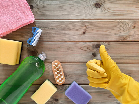Thumb up against background of kitchen utensils.  Top view. Hand in yellow glove shows like, next to it lie various cleaning items on wooden table. Flat lay. Perfect cleaning. Routine work in kitchen.
