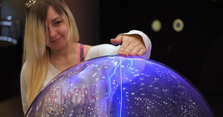 A woman runs her hand over a large ball of electrical discharges. Lightning strikes the woman's...