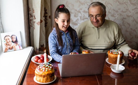 Grandfather And Granddaughter Are Talking Via Video Link To Their Friends. Decorated Table With Colorful Eggs And Cake. Chatting During The COVID Pandemic And The Easter Holidays