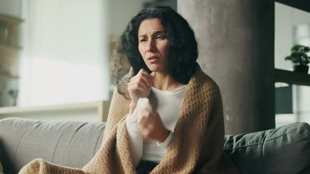 Sick hispanic young woman sitting on sofa under the warm blanket coughing sneezing and having severe headache temperature at home Unhealthy female getting flu virus symptom Cold and fever concept
