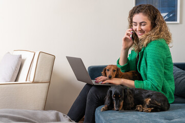 One blond woman wearing green outfit sitting on the couch working on the laptop and two Dachshund dogs 
