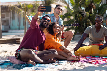 Happy diverse friends taking selfie and playing guitar at beach