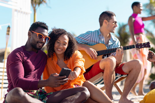 Happy diverse friends using smartphone and playing guitar at beach