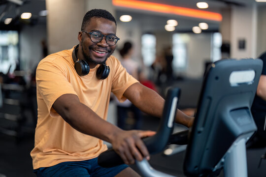 Portrait of satisfied male gym member with headphones around his neck looking at camera with hands on handlebars of stationary exercise bike cycling machine at gym.