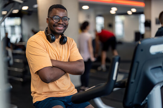 Portrait of smiling confident sportsman athlete with headphones around his neck looking at camera with arms crossed sitting on stationary exercise bike cycling machine at gym fitness center.