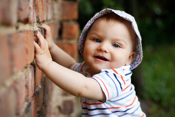 Cute 10-month-old baby boy wearing a hat and a striped T-shirt touching an old red brick wall in...