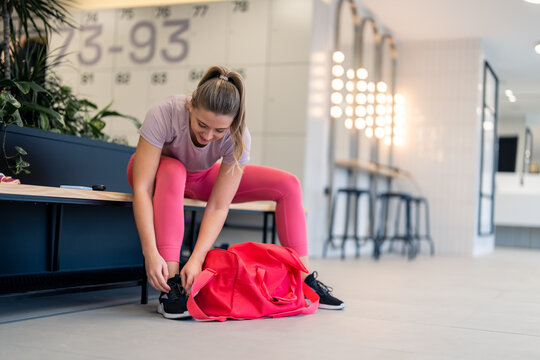 Young athletic sports woman with tied up hair wearing sportswear t-shirt and leggings sitting on bench in gym's locker room tying shoelaces on her sneakers while preparing for sports training session.