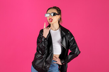 Cool biker girl wearing a leather jacket and jeans, gesturing sign of the horns. Isolated on pink background.