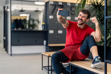 Handsome confident fit sports man sitting on bench in modern locker room at gym using smart mobile phone for video call, showing how happy and motivated he is for upcoming workout training session.