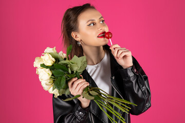 Cool biker girl wearing a leather jacket and jeans posing with a bouquet of white roses and lollipop. Isolated on pink background.