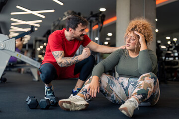 Supportive male professional gym fitness trainer being proud of his client active plus-size young woman with curly hair who is feeling exhausted and tired after hard workout individual training at gym