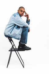 young and handsome Afro-American man in cool denim clothes sitting on the back of the chair and looking serious, studio shot vertical. High quality photo