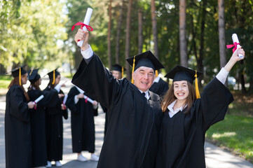 A group of graduates in robes outdoors. An elderly man and a young woman congratulate each other on...