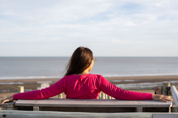 young woman sitting in front of the sea on a wooden seat with her arms open in relaxation mode