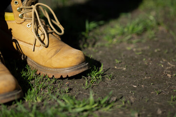 Sand-colored old farm male boots on garden soil. Man stands in garden on lawn with grass breaking through ground. Preparing for spring sowing season. Autumn. Farmer. Worker in safety shoes. Copyspace
