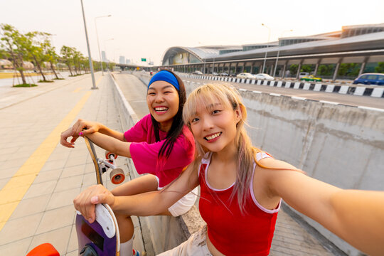 Asian woman friends using mobile phone taking selfie together during skating on longboard skate in the city. Stylish girl have fun outdoor lifestyle skateboarding on city street on summer vacation.