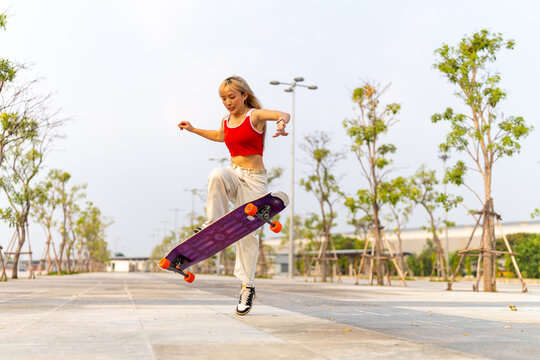 Cool Asian girl skating on longboard skate at park on summer holiday vacation. Stylish woman having fun urban outdoor active lifestyle practicing extreme sports skateboarding on city street at sunset.