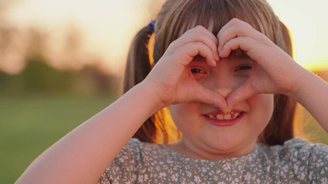 Cute little girl holding her hands in shape of heart in park. Concept of childrens love for family. Healthy child, play. Child folded his hands in form of heart outdoors, Portrait, kind smile of child