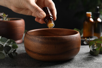 Woman dripping eucalyptus essential oil from bottle into bowl at light grey table, closeup