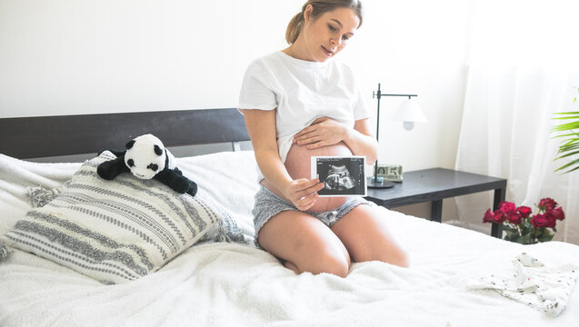 Pregnant woman sitting on a bed holding an X-ray image of her baby.The concept of pregnancy, motherhood and prenatal care. Mom with a new life.
