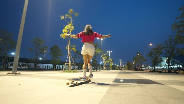 Cool Asian girl skating on longboard skate at park on summer holiday vacation. Stylish woman friends having fun urban outdoor active lifestyle practicing skateboarding together on city street at night