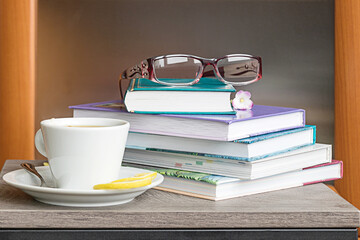 On the table are a stack of books for reading, women's glasses for vision, a cup of hot tea for a pleasant pastime.