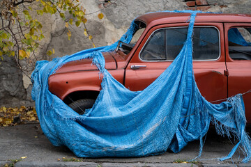 Retro old abandoned red car with broken glass covered with torn blue awning to prevent metal...