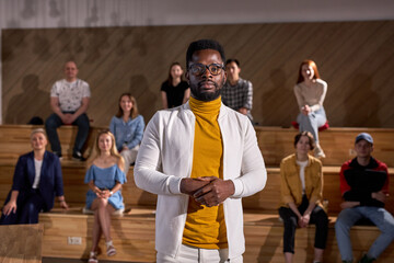 Portrait Of Handsome African American Lecturer In White Jacket Looking At Camera Posing Confidently, During Lecture Presentation To Group Of People In The Background, Lesson. Education. Copy Space