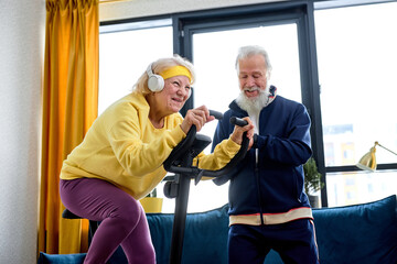 elderly lady training on stationary bicycle at home, with personal trainer gray haired man. Senior adult doing workout and physical exercise while using cardio bike machine for wellness and endurance