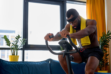 Young black fitness man training on a stationary bike, exercise at home alone. handsome male riding...