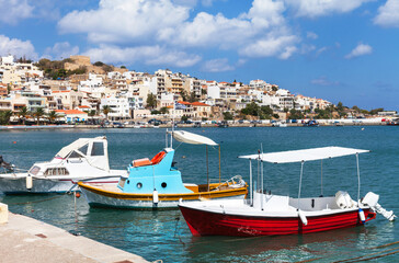 Fototapeta na wymiar Greece. View of Sitia town from Marina with traditional Greek pleasure bright boats against backdrop of blue sea bay and beautiful white houses on hill topped by ancient Kazarma fortress on sunny day
