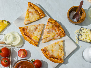 Top view shot sliced triangle pieces of delicious tasty homemade Hawaiian traditional Italian crust thin crispy ham and pineapple pizza placed on dish with ingredients tomatoes cheese salt on table