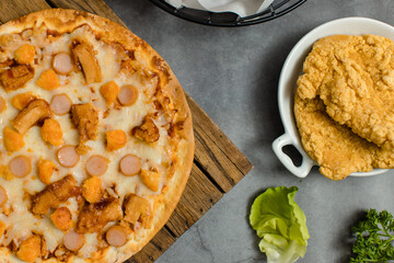 Top view shot of delicious tasty juicy sliced sausage cheesy pizza placed on wooden board around with other fast food crispy fried chicken, seasoning ingredients and fresh raw organic vegetables