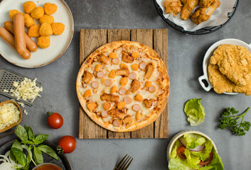 Top view shot of delicious tasty juicy sliced sausage cheesy pizza placed on wooden board around with other fast food crispy fried chicken, seasoning ingredients and fresh raw organic vegetables