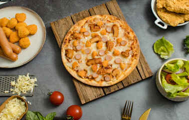 Top view shot of delicious tasty juicy sliced sausage cheesy pizza placed on wooden board around with other fast food crispy fried chicken, seasoning ingredients and fresh raw organic vegetables