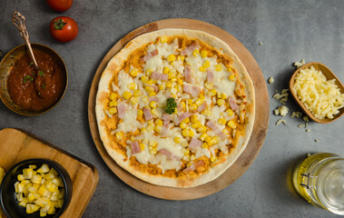 Top view closeup shot of delicious tasty juicy thin crispy cheesy corn and ham Italian pizza placed on wooden board on party table around with other ingredients, sauce, olive oil, corn and tomatoes.