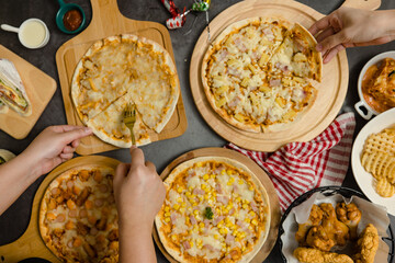 Top view shot of various kinds of delicious tasty juicy cheesy pizza placed on wooden board on party table around with other fast food while unrecognizable people picking up eating dinner together