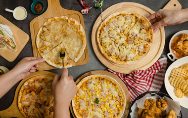 Top view shot of various kinds of delicious tasty juicy cheesy pizza placed on wooden board on party table around with other fast food while unrecognizable people picking up eating dinner together