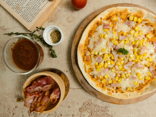 Top view closeup shot of delicious tasty juicy thin crispy cheesy corn and ham Italian pizza placed on wooden board on party table around with other ingredients, sauce, olive oil, bacon and tomatoes