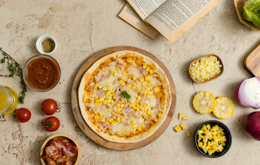Top view shot of delicious tasty juicy cheesy corn and ham Italian pizza placed on wooden board on party table around with other ingredients, sauce, olive oil, bacon, tomatoes, cheese and vegetables