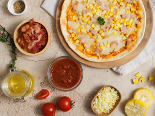 Top view shot of delicious tasty juicy cheesy corn and ham Italian pizza placed on wooden board on party table around with other ingredients, sauce, olive oil, bacon, tomatoes, cheese and vegetables