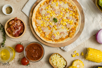 Top view shot of delicious tasty juicy cheesy corn and ham Italian pizza placed on wooden board on party table around with other ingredients, sauce, olive oil, bacon, tomatoes, cheese and vegetables