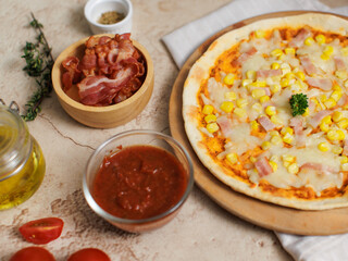 Top view closeup shot of delicious tasty juicy thin crispy cheesy corn and ham Italian pizza placed on wooden board on party table around with other ingredients, sauce, olive oil, bacon and tomatoes