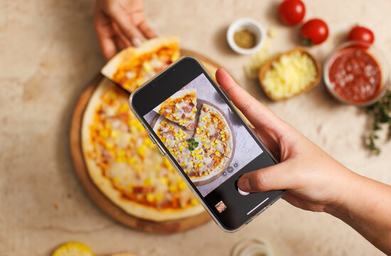 Unrecognizable person using smartphone taking photo top view shot of delicious tasty juicy corn and ham cheesy pizza placed on wooden board on table while people hand picking up piece from whole