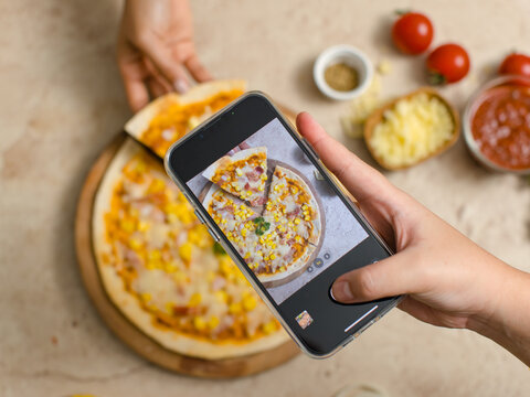 Unrecognizable person using smartphone taking photo top view shot of delicious tasty juicy corn and ham cheesy pizza placed on wooden board on table while people hand picking up piece from whole