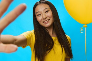 Asian woman holding a yellow balloon on a blue background in a yellow T-shirt smiling with teeth,...